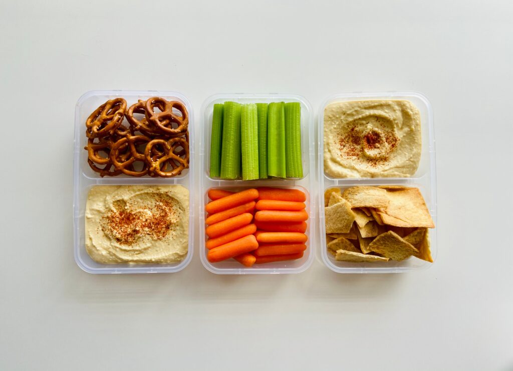 A portioned container holding pretzels, hummus, celery, carrots, and crackers.