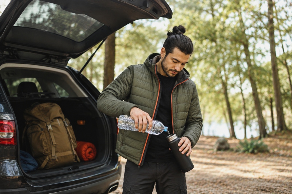 A man standing outside at the back of his car that has the trunk open. Inside the care is a backpack and a rolled up sleeping bag. The man is filling his refillable water bottle with water from a plastic bottle.